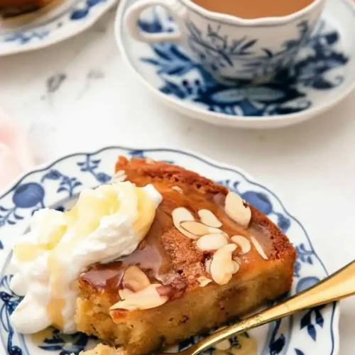Une part appétissante de gâteau aux amandes et à la crème fouettée, arrosée de miel, servie avec une tasse de café dans une vaisselle délicate à motifs bleus, invitant à savourer un moment gourmand avec ce gateau au miel