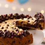 Un gateau cookie festif pour célébrer : Une tranche est soulevée d'un gateau cookie décoré de ganache au chocolat et de vermicelles colorés. Des ballons en arrière-plan suggèrent une fête, parfait pour partager ce délicieux gateau cookie.