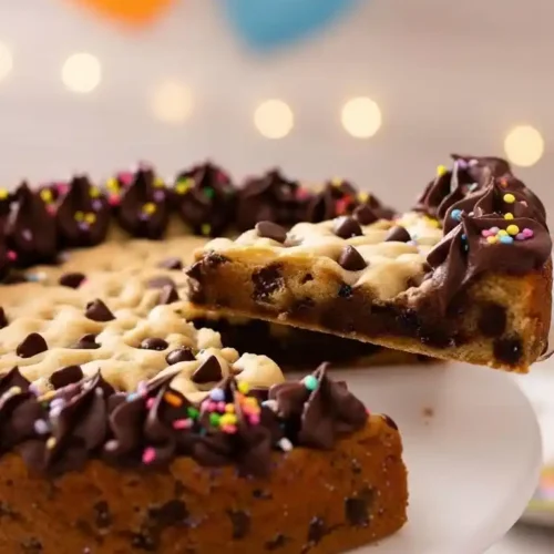 Un gateau cookie festif pour célébrer : Une tranche est soulevée d'un gateau cookie décoré de ganache au chocolat et de vermicelles colorés. Des ballons en arrière-plan suggèrent une fête, parfait pour partager ce délicieux gateau cookie.