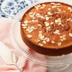 Un délicieux gâteau doré aux amandes, servi sur un présentoir à gâteau en verre avec un fond de livre ouvert et d'assiette décorative, évoquant la douceur et le plaisir d'un gateau au miel parfait.
