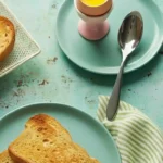 Photo d'un petit déjeuner gourmand composé d'un œuf à la coque présenté dans un coquetier rose, accompagné de toasts dorés disposés sur des assiettes bleues, avec une cuillère et une serviette rayée verte et blanche. Une composition appétissante invitant à savourer un moment de détente et de plaisir matinal.