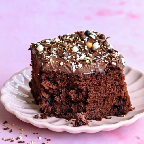 Un appétissant morceau de gâteau des écoliers est présenté sur une assiette rose. Le gâteau au chocolat est nappé d'un glaçage chocolaté et décoré d'une généreuse quantité de vermicelles colorés. Quelques miettes et vermicelles éparpillés autour de l'assiette ajoutent une touche gourmande à la scène. Le fond rose accentue les couleurs du dessert.
