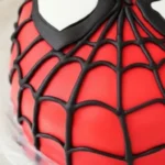 Gâteau Spiderman impressionnant: un gâteau détaillé en forme de tête de Spiderman, avec un glaçage rouge, une toile d'araignée noire et des yeux blancs distinctifs, idéal pour une fête d'anniversaire.