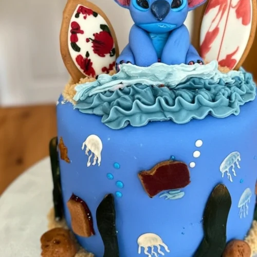 Un adorable gâteau Stitch décoré sur le thème de l'océan, avec une figurine de Stitch en pâte à sucre, des vagues en glaçage bleu, des méduses et des algues, évoquant une ambiance hawaïenne. Des biscuits en forme de planches de surf complètent le décor.