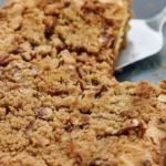 Un gateau petit dejeuner aux noix, doré et croustillant, est présenté dans un plat en verre. Une part est soulevée par une spatule, révélant une mie moelleuse et un crumble riche en saveurs. Idéal pour un réveil gourmand