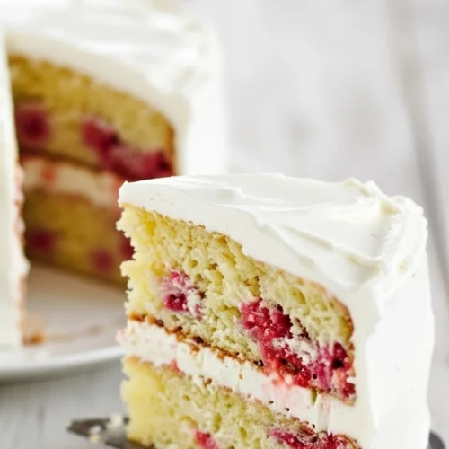 Gros plan d'une part de gateau framboise à deux étages, avec un glaçage blanc crémeux et des framboises fraîches, reposant sur une pelle à tarte en métal.