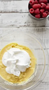 Préparation du gateau framboise : vue de dessus d'un bol contenant de la pâte à gâteau et des blancs d'œufs montés en neige, avec une tasse de framboises fraîches à côté.
