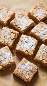 Un assortiment de carrés de gateau aux pommes saupoudrés de sucre glace, disposés sur du papier sulfurisé marron. Chaque part de gateau aux pommes a une texture moelleuse et contient des morceaux de pomme visibles.