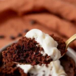 Gros plan tentant d'une portion de gateau aux chocolat au micro onde, présentée sur une cuillère dorée et nappée de crème fouettée onctueuse. Les détails de la mie aérée et des inclusions de chocolat noir invitent à la dégustation rapide de ce dessert express.