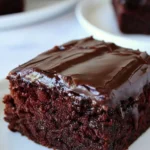 Gros plan sur une part carrée de gâteau chocolat rapide et moelleux, nappée d'un glaçage brillant qui coule sur le côté, servie sur une assiette blanche.