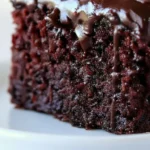 Gros plan vertical sur une part de gâteau chocolat rapide ultra moelleux, couvert d'un glaçage riche et brillant qui coule sur le bord, posé sur une assiette blanche.