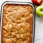 Vue de dessus d'un gateau pomme doré, cuit dans un plat rectangulaire en verre. La texture du gâteau est légèrement craquelée, avec des morceaux de pommes visibles à la surface. Une pomme rouge et une pomme verte sont placées à côté du plat, sur une surface claire. Un délicieux gateau pomme fait maison prêt à être dégusté.