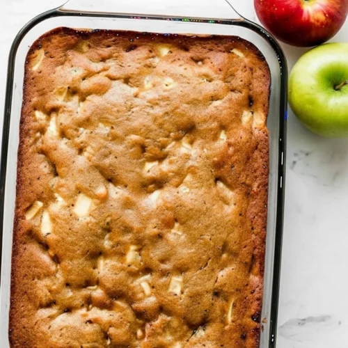 Vue de dessus d'un gateau pomme doré, cuit dans un plat rectangulaire en verre. La texture du gâteau est légèrement craquelée, avec des morceaux de pommes visibles à la surface. Une pomme rouge et une pomme verte sont placées à côté du plat, sur une surface claire. Un délicieux gateau pomme fait maison prêt à être dégusté.