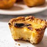 Une photographie appétissante d'un pastel de nata, un délicieux gâteau portugais, avec une bouchée enlevée. On peut voir sa garniture à la crème riche et son extérieur feuilleté, croustillant et doré. D'autres pasteis de nata, flous en arrière-plan, suggèrent une délicieuse sélection de ce classique gâteau portugais.