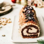 Gros plan sur une bûche de noël praliné croustillant roulée à la génoise chocolat, garnie de copeaux de chocolat sur le dessus et présentée dans une ambiance festive avec des boules rouges et dorées
