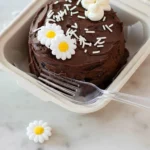 Vue verticale du résultat final de la recette bento cake : un mini gâteau au chocolat présenté dans sa boîte à emporter, décoré de marguerites en sucre et accompagné d'une fourchette en plastique prête à la dégustation.