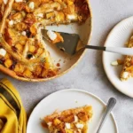 Vue de dessus d'une part de tarte butternut feta servie sur une petite assiette blanche, avec le reste du plat entamé en arrière-plan. On y voit des cubes de courge rôtie, des morceaux de feta et une pâte dorée croustillante, le tout accompagné d'une serviette jaune.