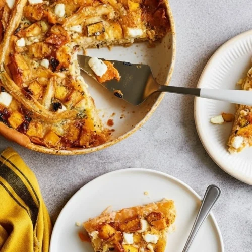 Vue de dessus d'une part de tarte butternut feta servie sur une petite assiette blanche, avec le reste du plat entamé en arrière-plan. On y voit des cubes de courge rôtie, des morceaux de feta et une pâte dorée croustillante, le tout accompagné d'une serviette jaune.