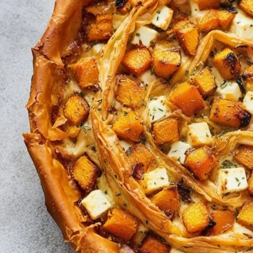 Gros plan détaillé sur une tarte butternut feta à la pâte croustillante et dorée, garnie de cubes de courge rôtie, de dés de fromage et d'herbes aromatiques.